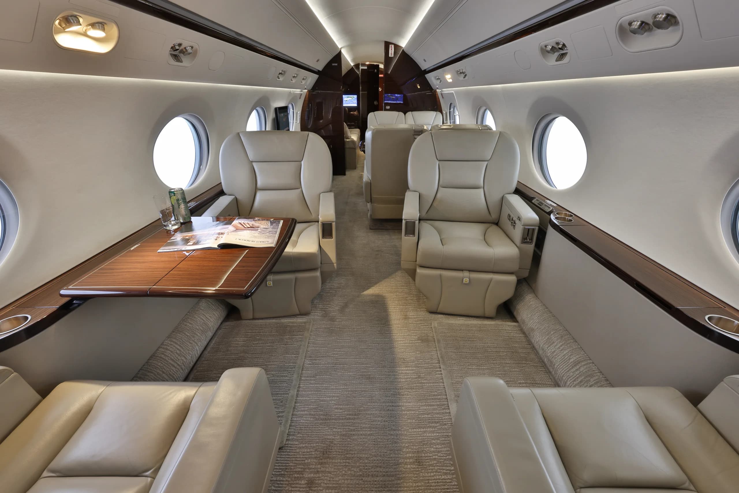 A luxurious aircraft cabin with beige leather seats, wooden tables, and large oval windows.
