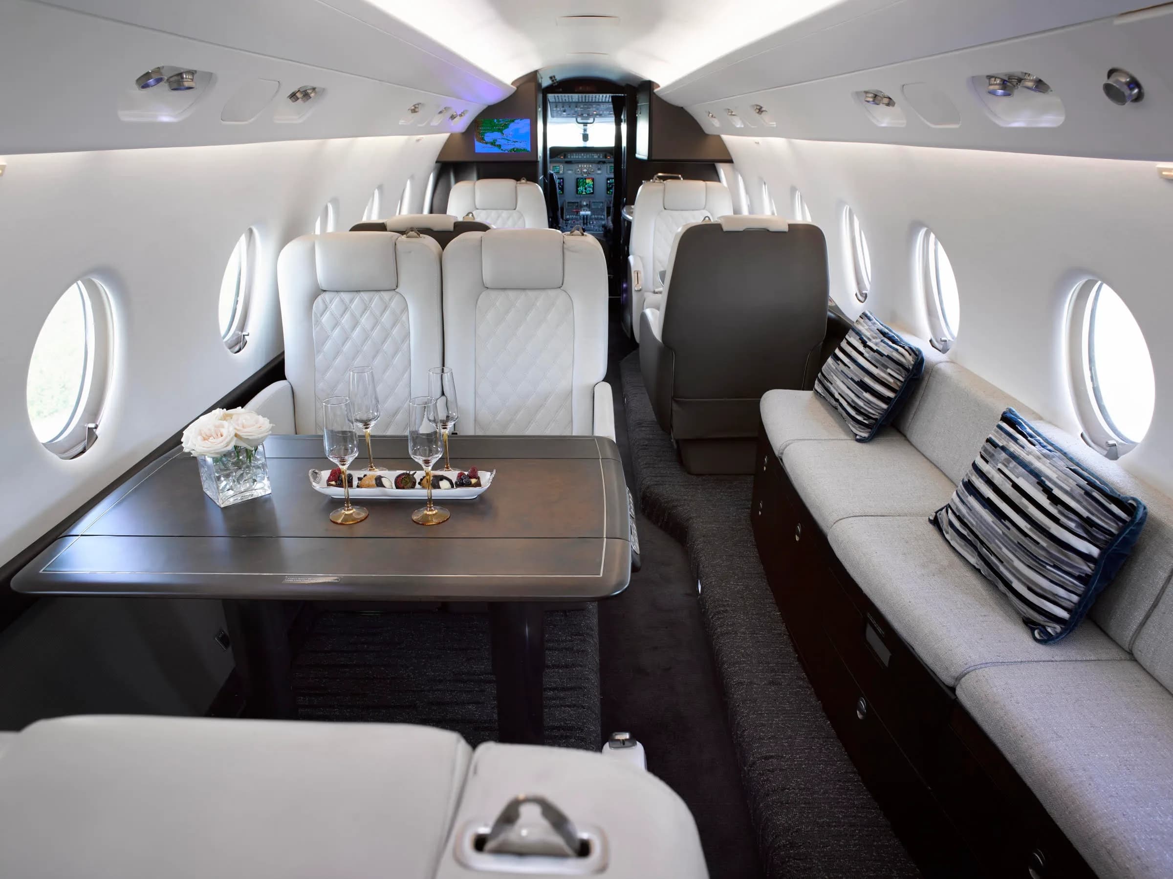 The luxurious interior of a private jet cabin features white quilted seats, a table set with drinks and snacks, and a view towards the cockpit.