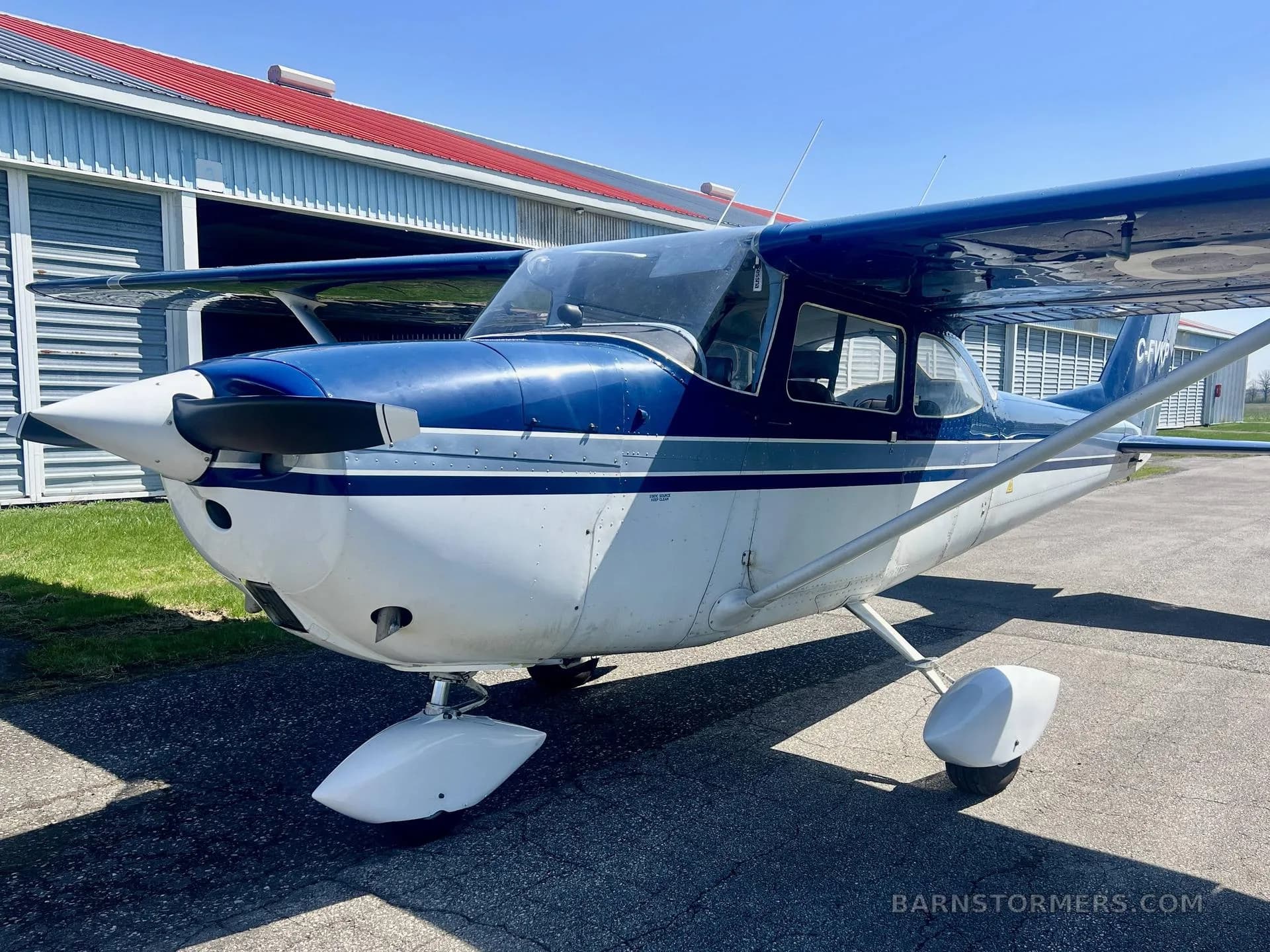 A blue and white 1967 Cessna 172H, registration C-FVKP, parked on an tarmac in front of a hangar, viewed from the front-right.