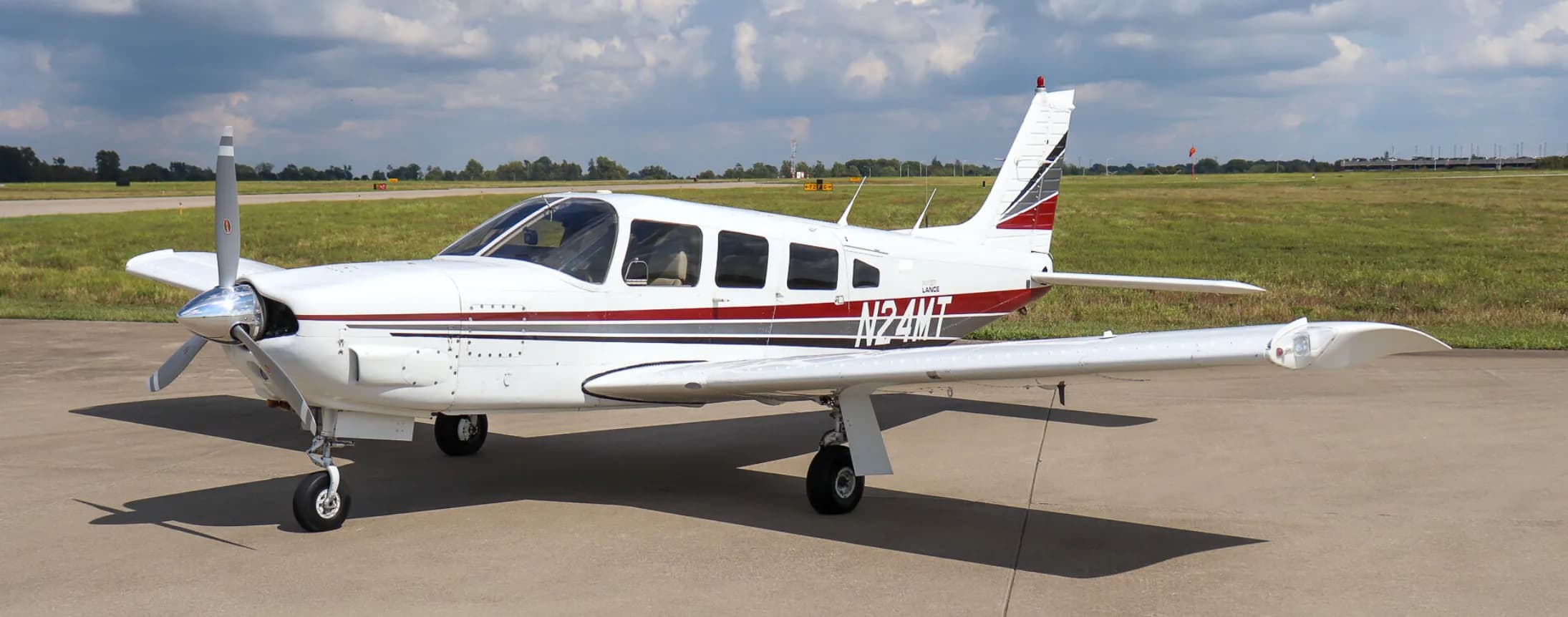 A white Piper Lance (N24MT) with red and grey stripes is parked on an airfield, viewed from the front-left.
