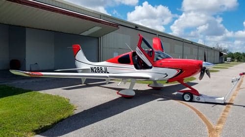 A red and white single-engine aircraft, N288JL, with its doors open, parked on an airfield tarmac next to a hangar.