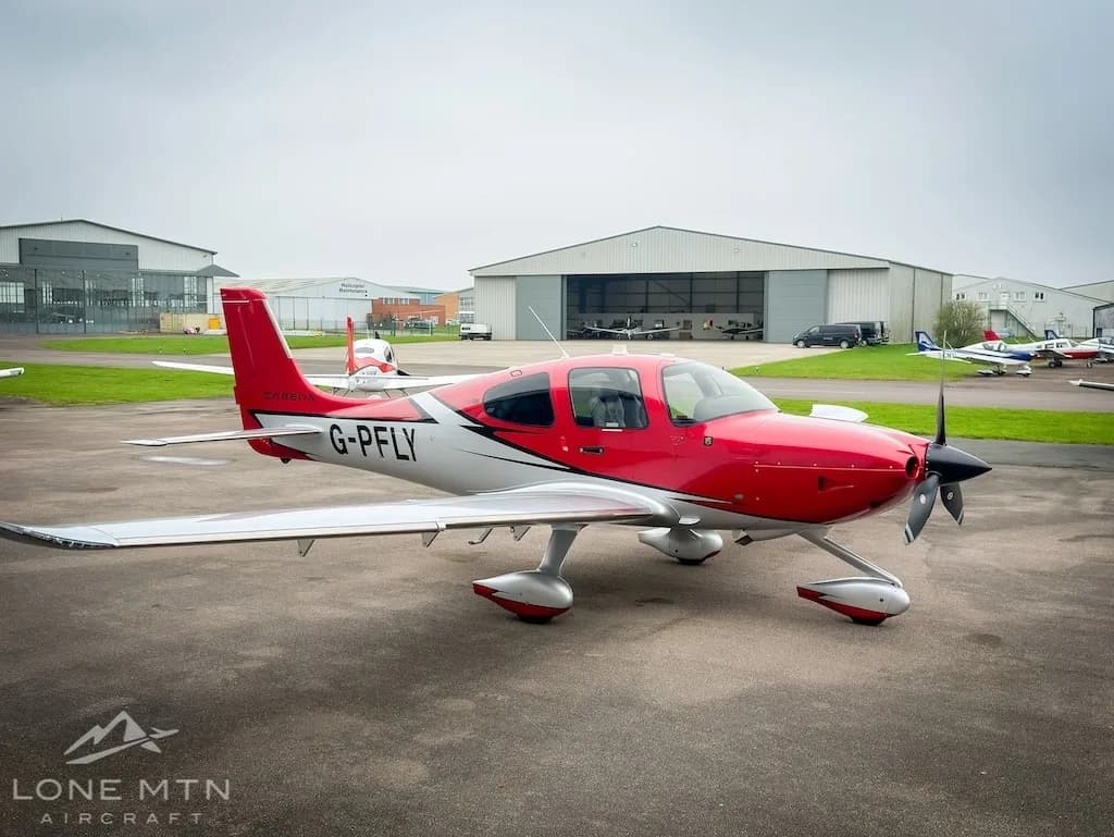 A red and white Cirrus SR22 aircraft (G-PFLY) parked on a tarmac with hangars and other aircraft in the background.