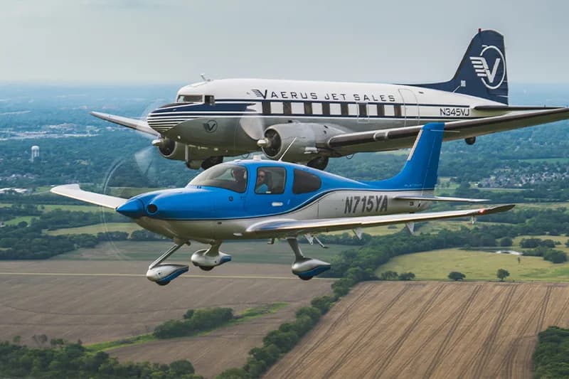 A Cirrus and a Douglas DC-3 fly in formation over a rural landscape.