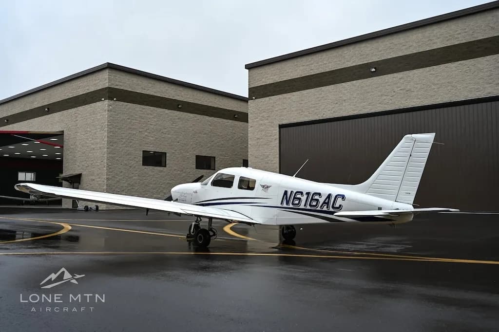 A white single-engine aircraft, N616AC, with blue stripes is parked on a wet tarmac next to hangars.