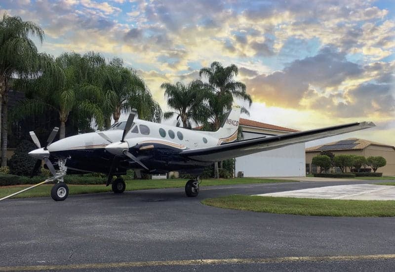 A Beechcraft King Air 90C aircraft, registration N94CD, parked on a paved surface with palm trees and a building in the background.