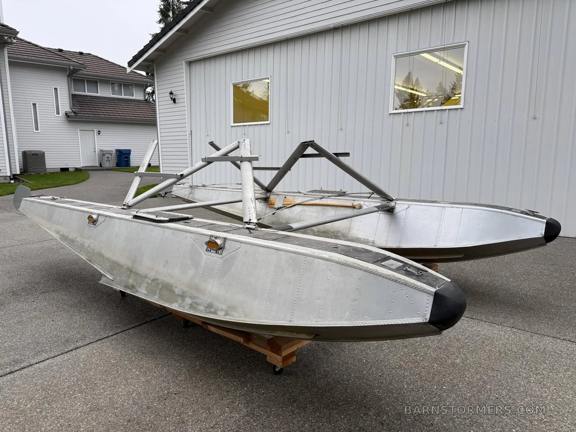 A pair of aluminum aircraft floats with mounting struts, viewed from a front-side angle, on a concrete driveway.