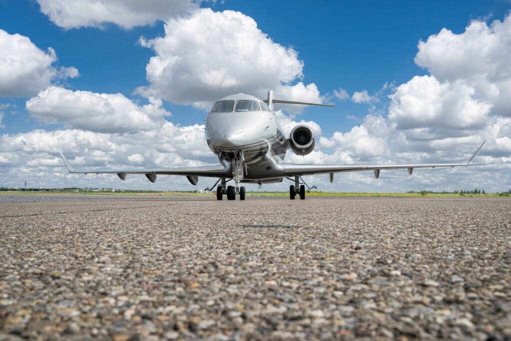 A silver and gray Challenger aircraft sits on the tarmac under a blue sky with clouds, facing forward.