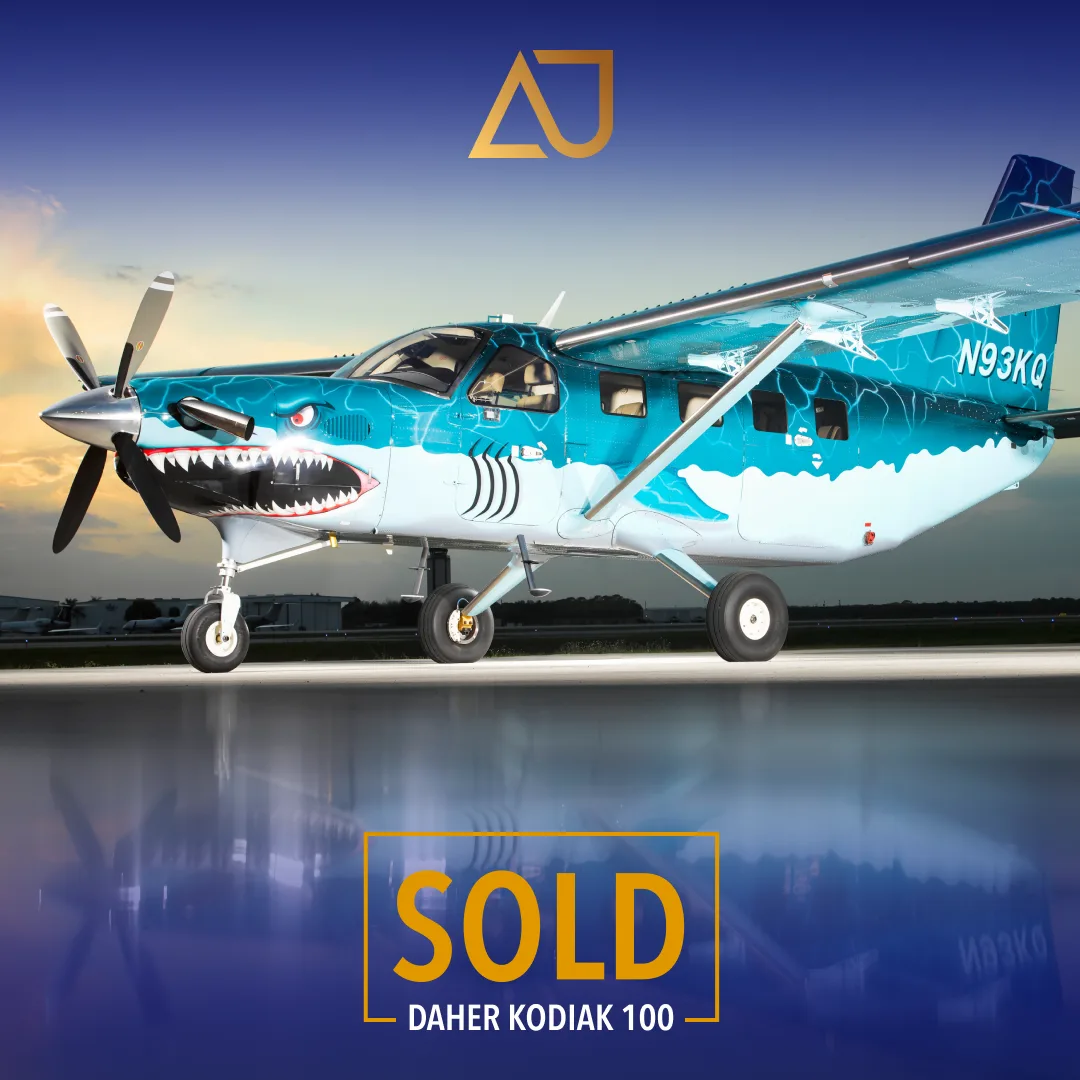 A Daher Kodiak 100 aircraft with a blue and white shark-mouth paint scheme, registration N93KQ, parked on a tarmac at sunset.