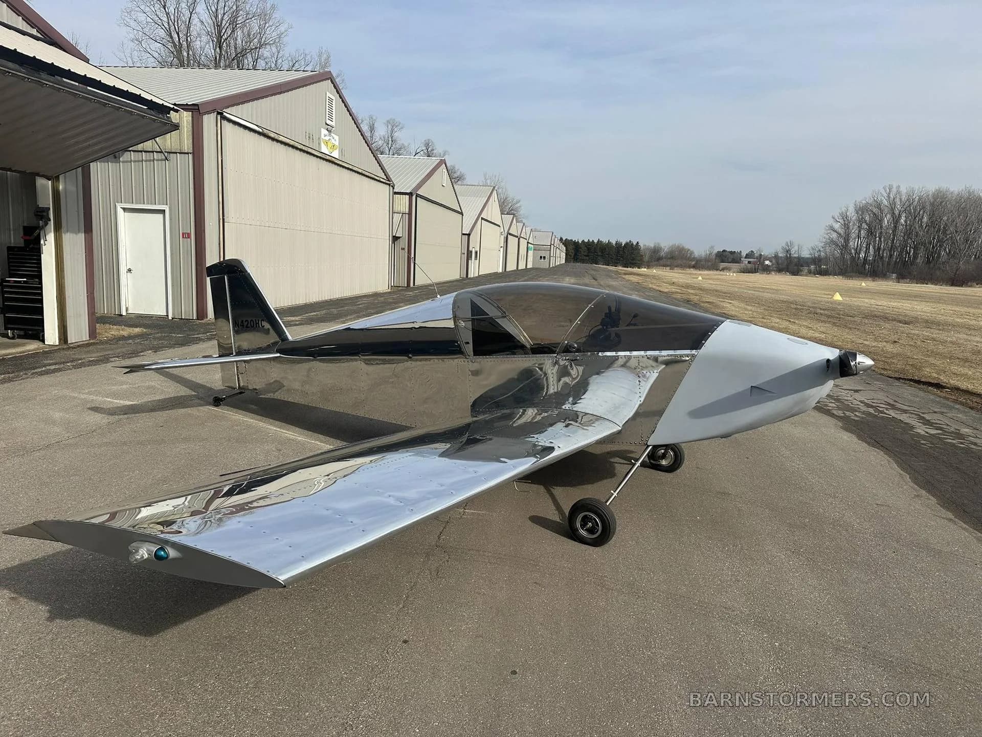 A shiny silver and black experimental aircraft, possibly a Sonex, parked on an asphalt taxiway next to hangars.