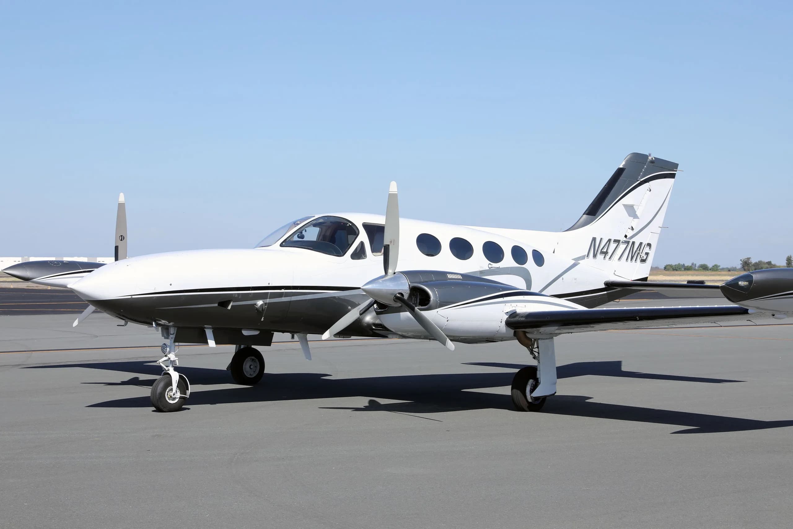 A white and grey twin-engine Cessna 414 Chancellor aircraft, N477MG, parked on an asphalt tarmac under a clear sky.