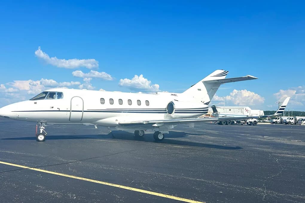 A white Hawker business jet with black stripes on the tarmac under a blue sky, with ground vehicles in the background.