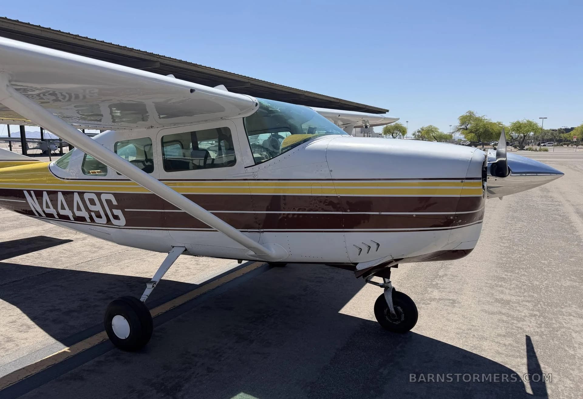 A front-side view of a white, yellow, and brown Cessna 182 aircraft with registration N4449G parked on an asphalt apron.