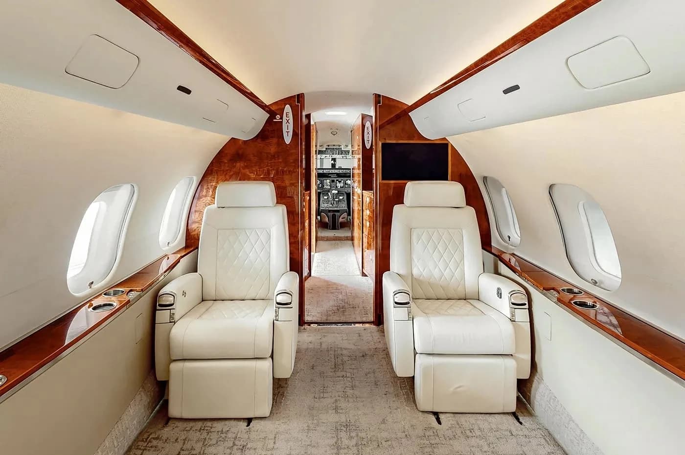 A view from the rear of the business jet cabin looking forward, showing two facing light-colored quilted seats and the entrance to the cockpit.