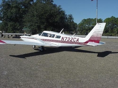 A Piper Apache aircraft with a white and maroon paint scheme is parked on the grass.
