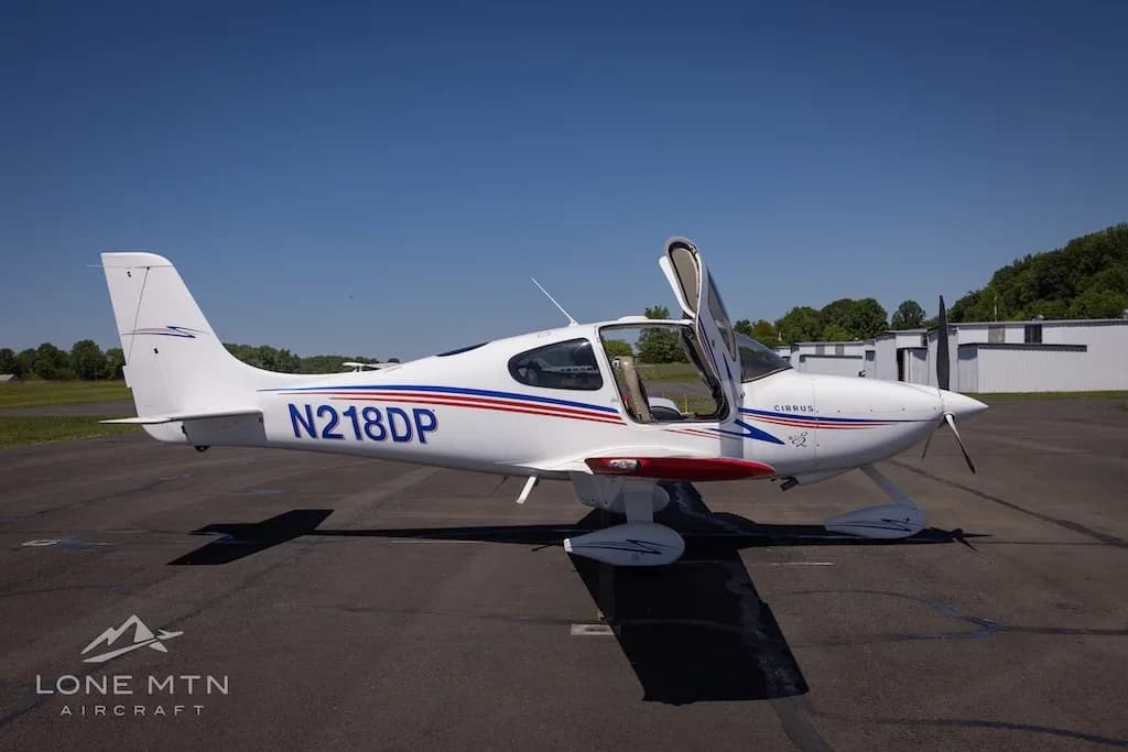 Side view of a white Cirrus aircraft (N218DP) with blue and red stripes and red wingtips, doors open, parked on an airfield.