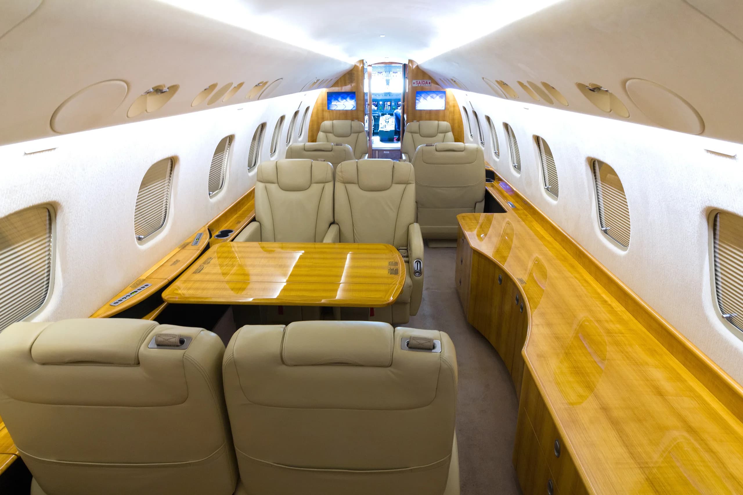 A wide view of a private jet cabin interior with multiple beige leather seats, polished wooden tables, and overhead lighting.