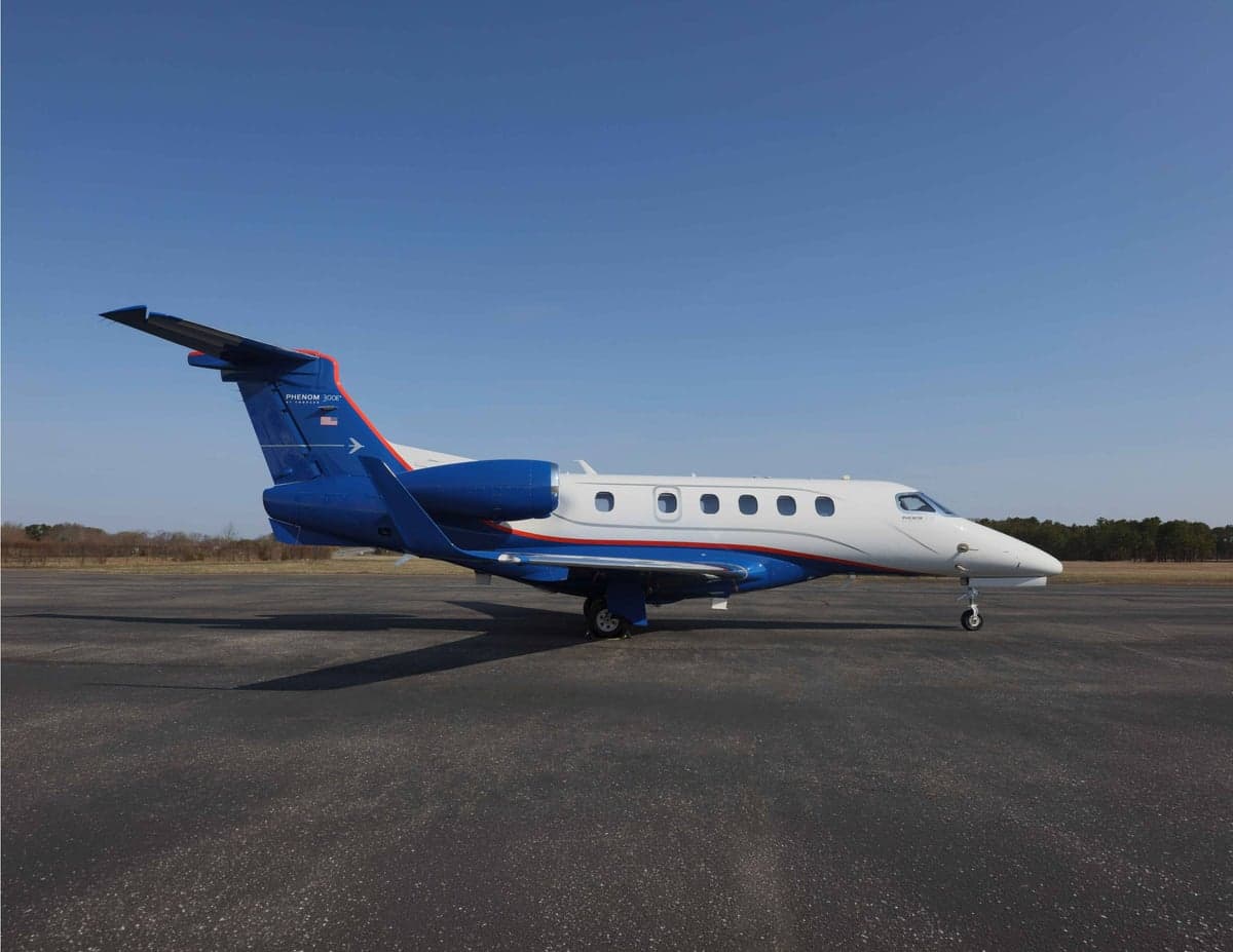 A Phenom 300E aircraft is parked on the tarmac with a blue and white paint scheme.