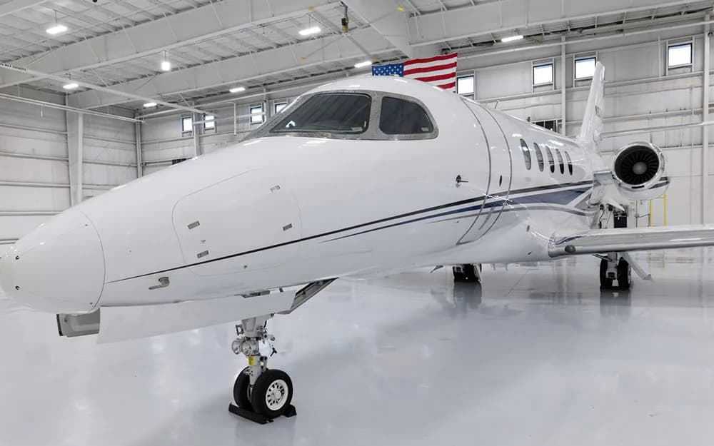 A front-quarter view of a white business jet with blue and black stripes, parked in a hangar with an American flag in the background.