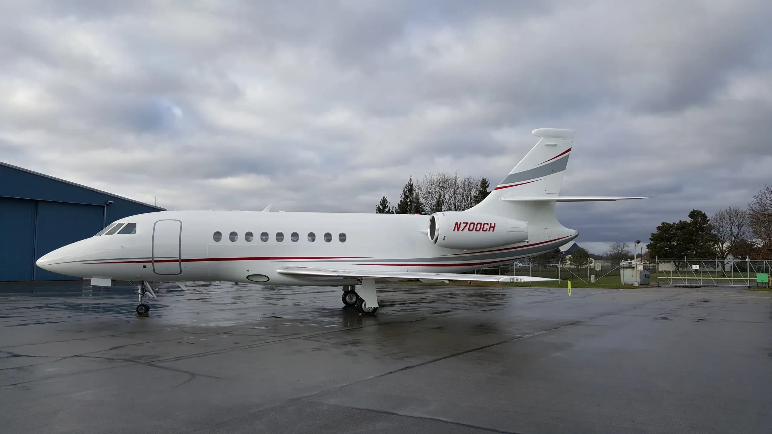 A white Dassault Falcon 2000 business jet, registration N700CH, with red and grey stripes, parked on a wet tarmac.
