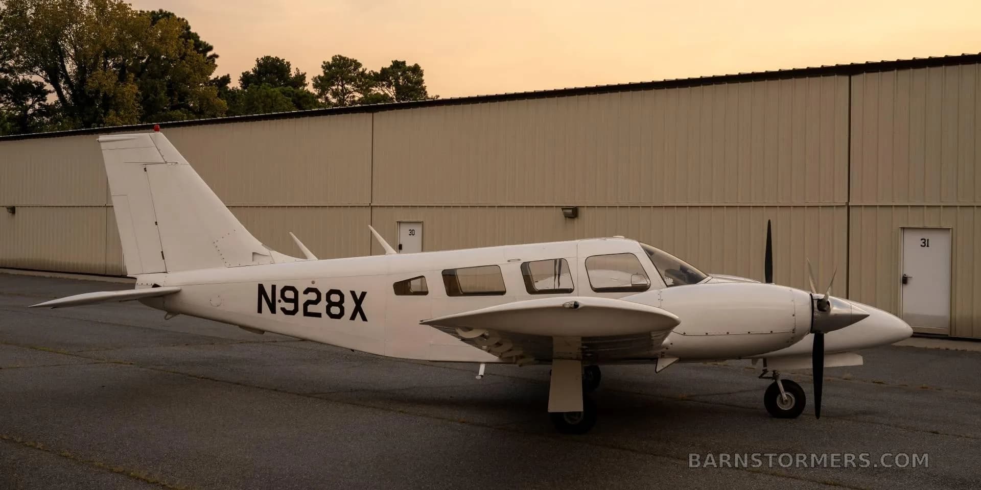 A white twin-engine propeller aircraft (N928X) is parked on an asphalt ramp with a hangar in the background at sunset.