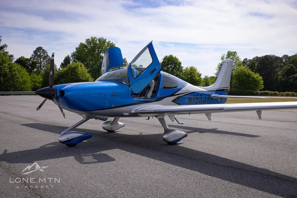 A blue and silver single-engine aircraft with open gull-wing doors is parked on an asphalt tarmac.