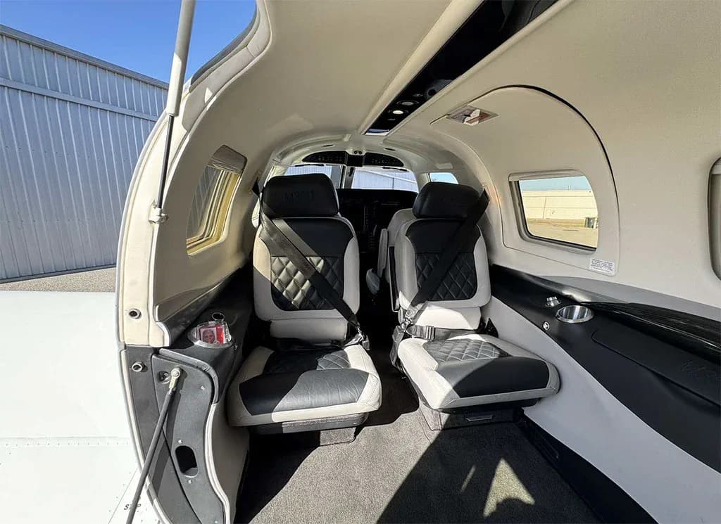 The aircraft's interior cabin features two rows of black and white quilted leather seats with seatbelts, looking towards the cockpit.
