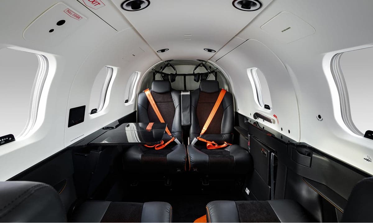 The aircraft cabin interior with black leather seats, orange seatbelts, and a small table.