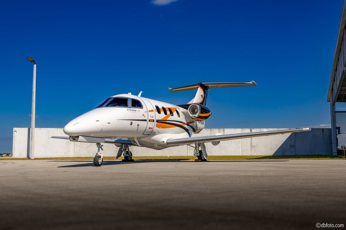 A Phenom 100 aircraft with a white, orange, and black paint scheme parked on the tarmac.