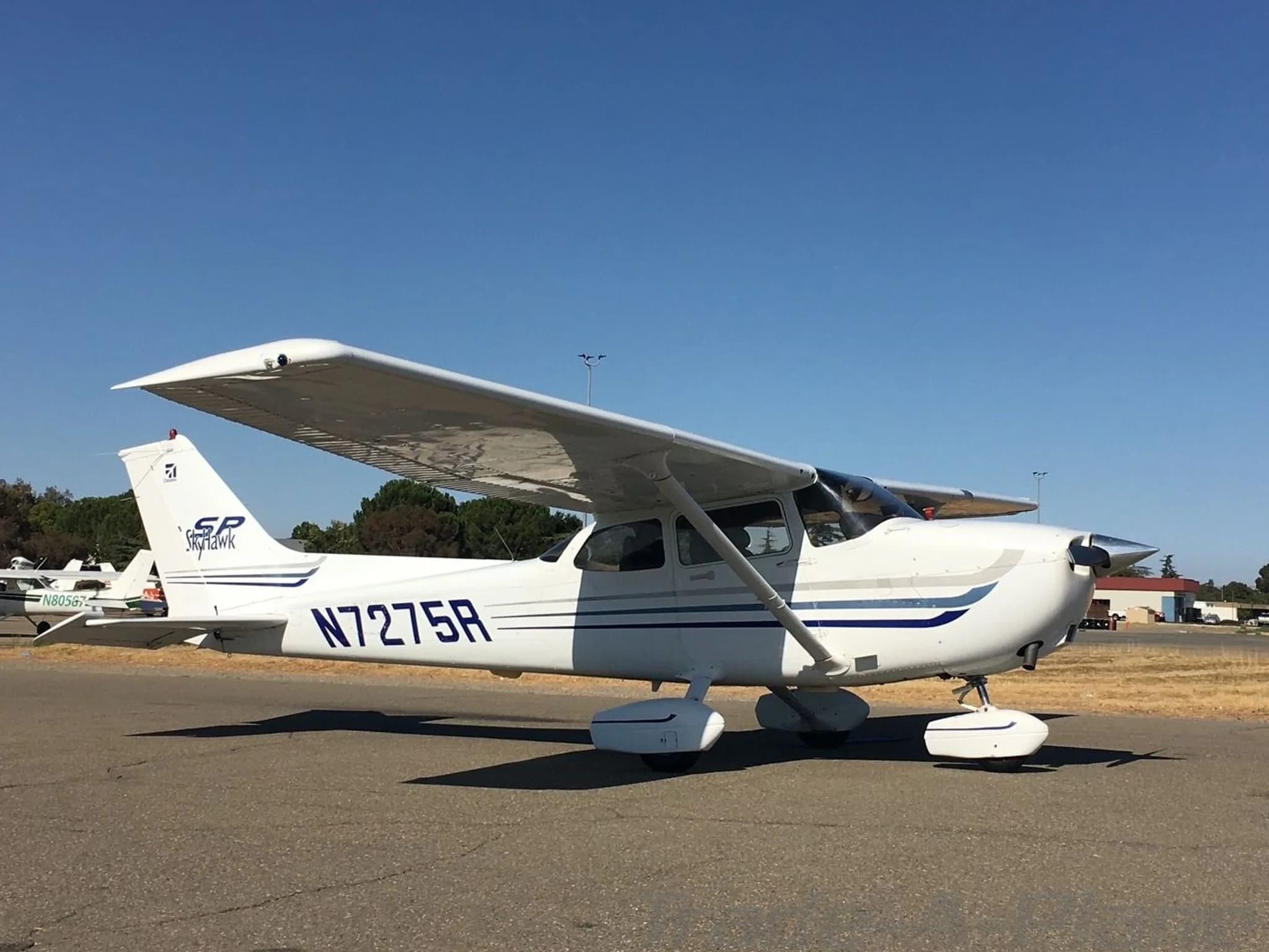 A white Cessna 172SP Skyhawk with blue stripes, registration N7275R, parked on an asphalt ramp with another aircraft in the background.