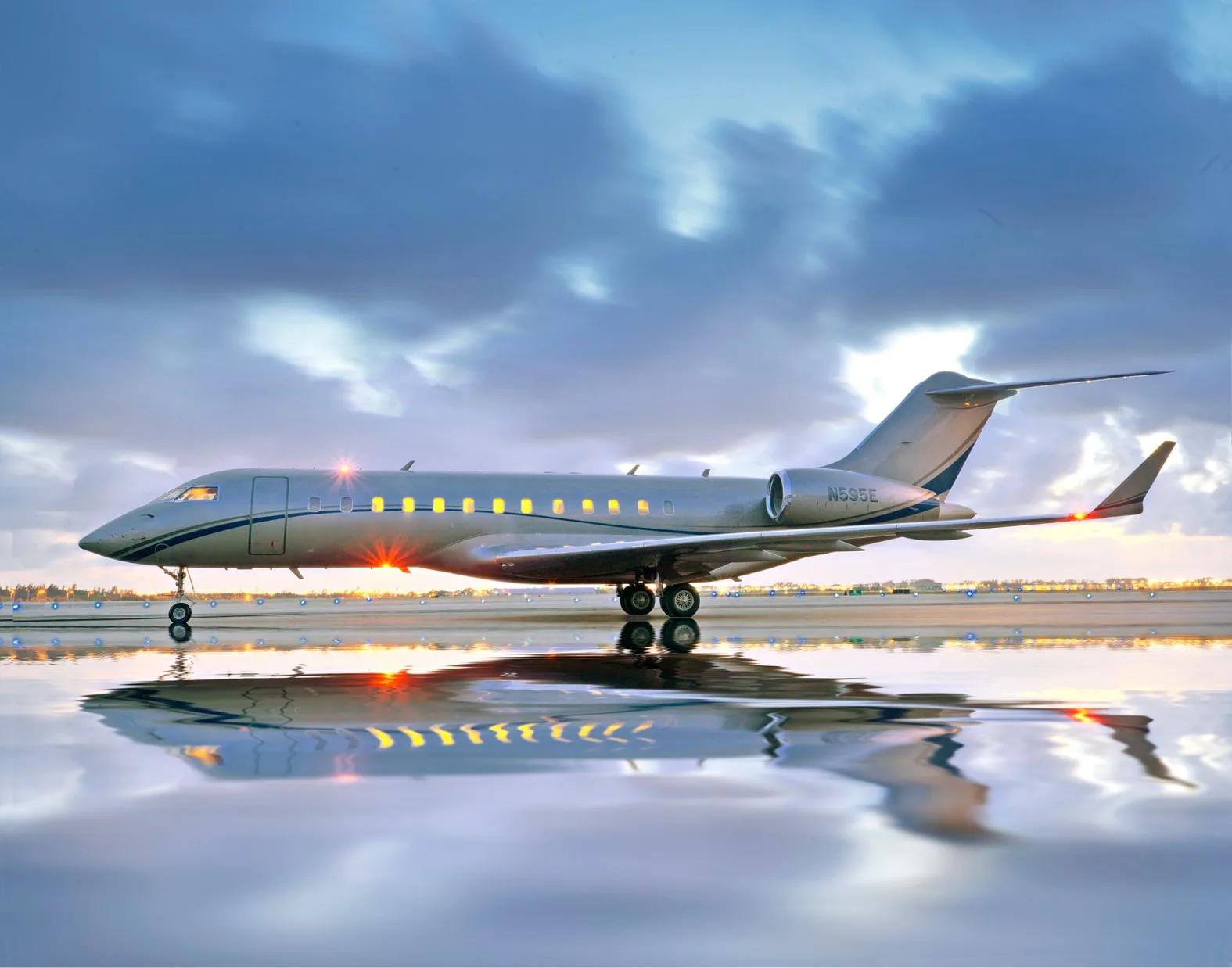 A silver Bombardier Global Express private jet (N99BE) with its lights on, reflected on a wet runway at dusk.