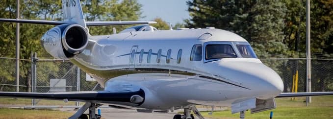 A white business jet with dark stripes is parked on an tarmac, seen from the front-right, with a fence and trees in the background.
