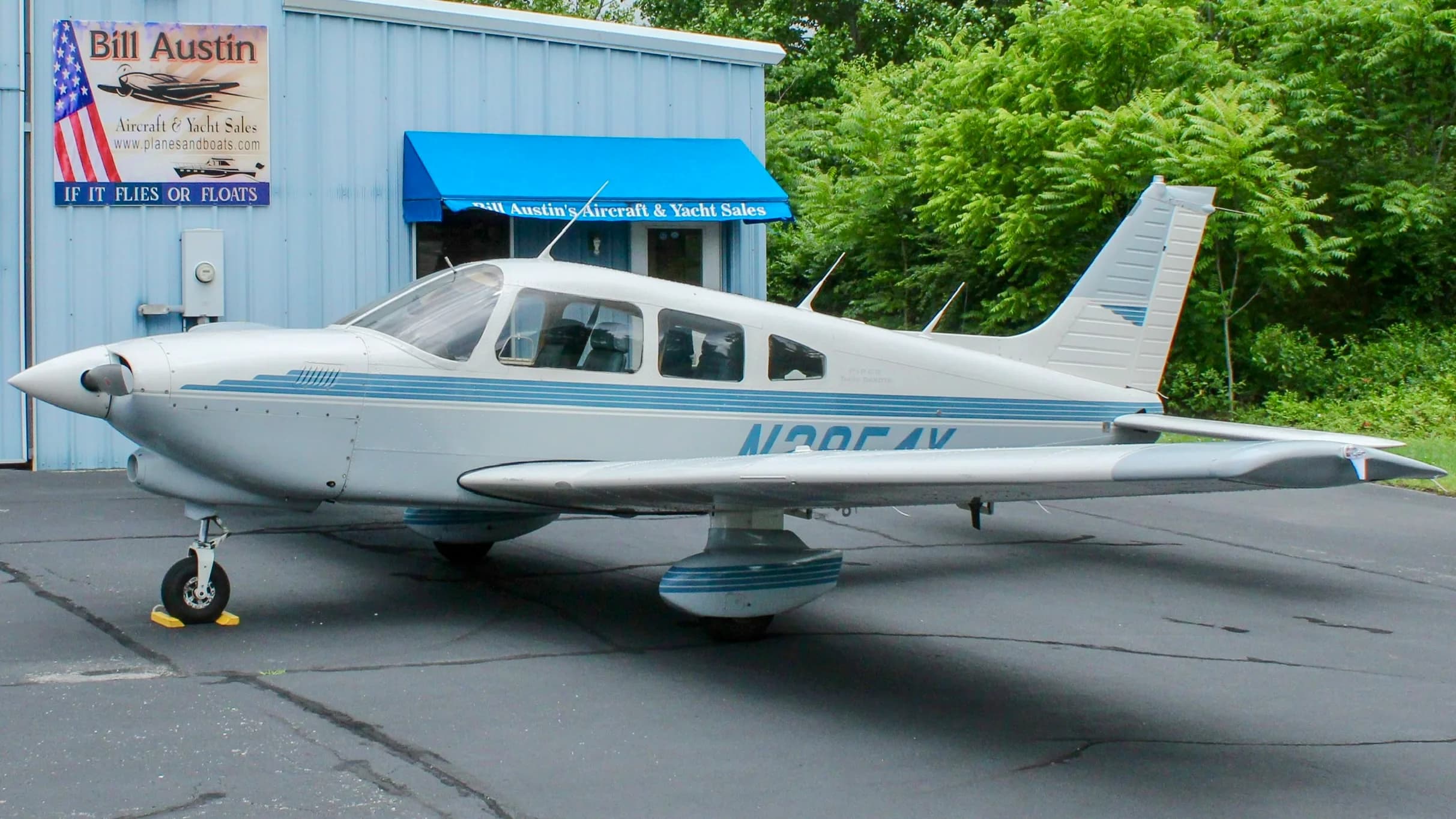 A white and blue Piper Turbo Dakota aircraft is parked on an asphalt lot in front of a light blue building.