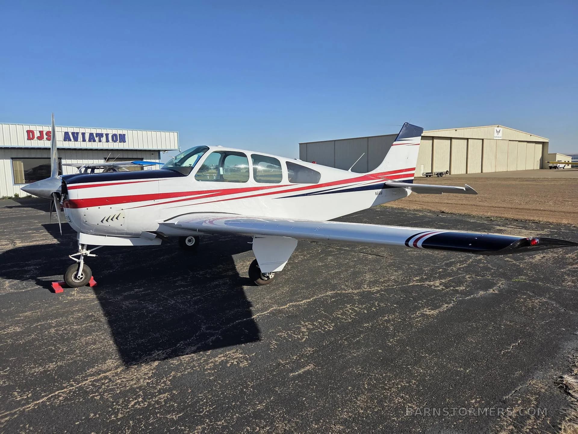 A white and red single-engine aircraft with dark wingtips is parked on an asphalt tarmac in front of a hangar labeled DJS AVIATION.