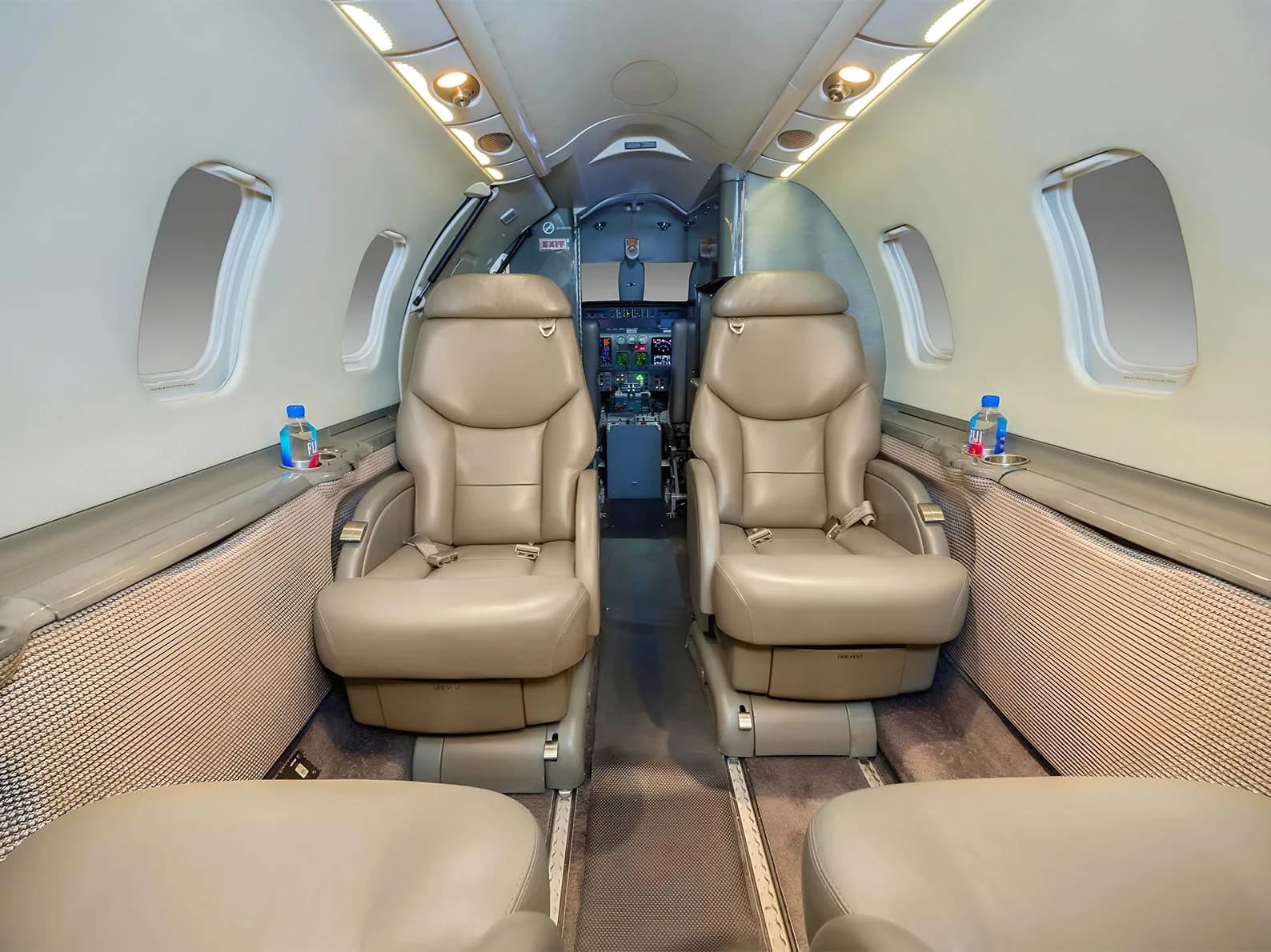 The forward cabin of a business jet with two beige leather seats facing forward and the cockpit visible in the background.