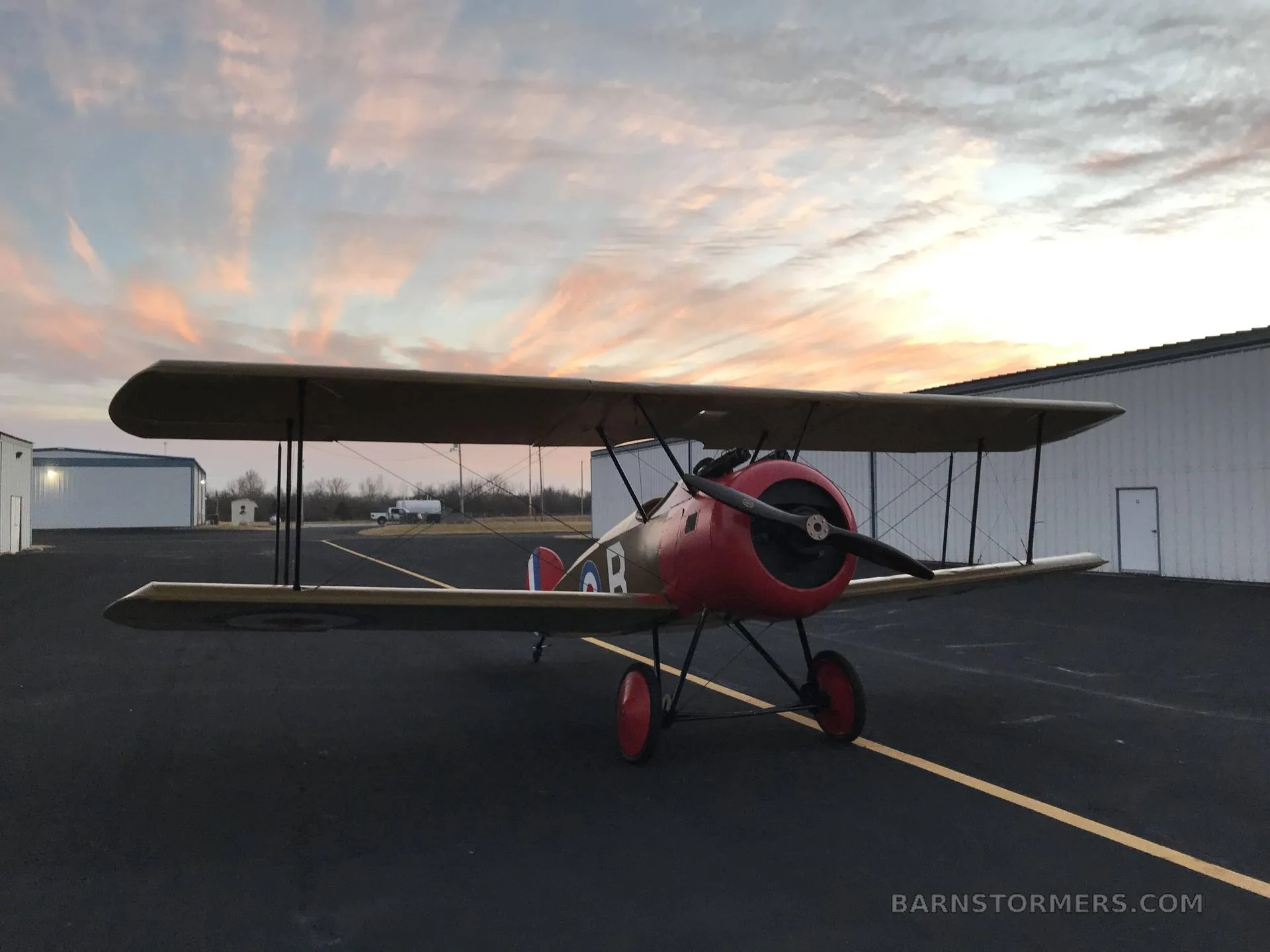 A tan and red biplane with British roundels is parked on an asphalt tarmac at sunset, with hangars in the background.