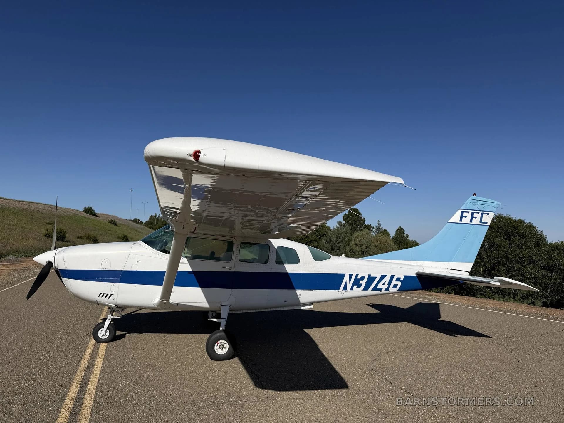 A white and blue Cessna 182 Skylane with registration N3746 parked on an asphalt surface under a clear blue sky.