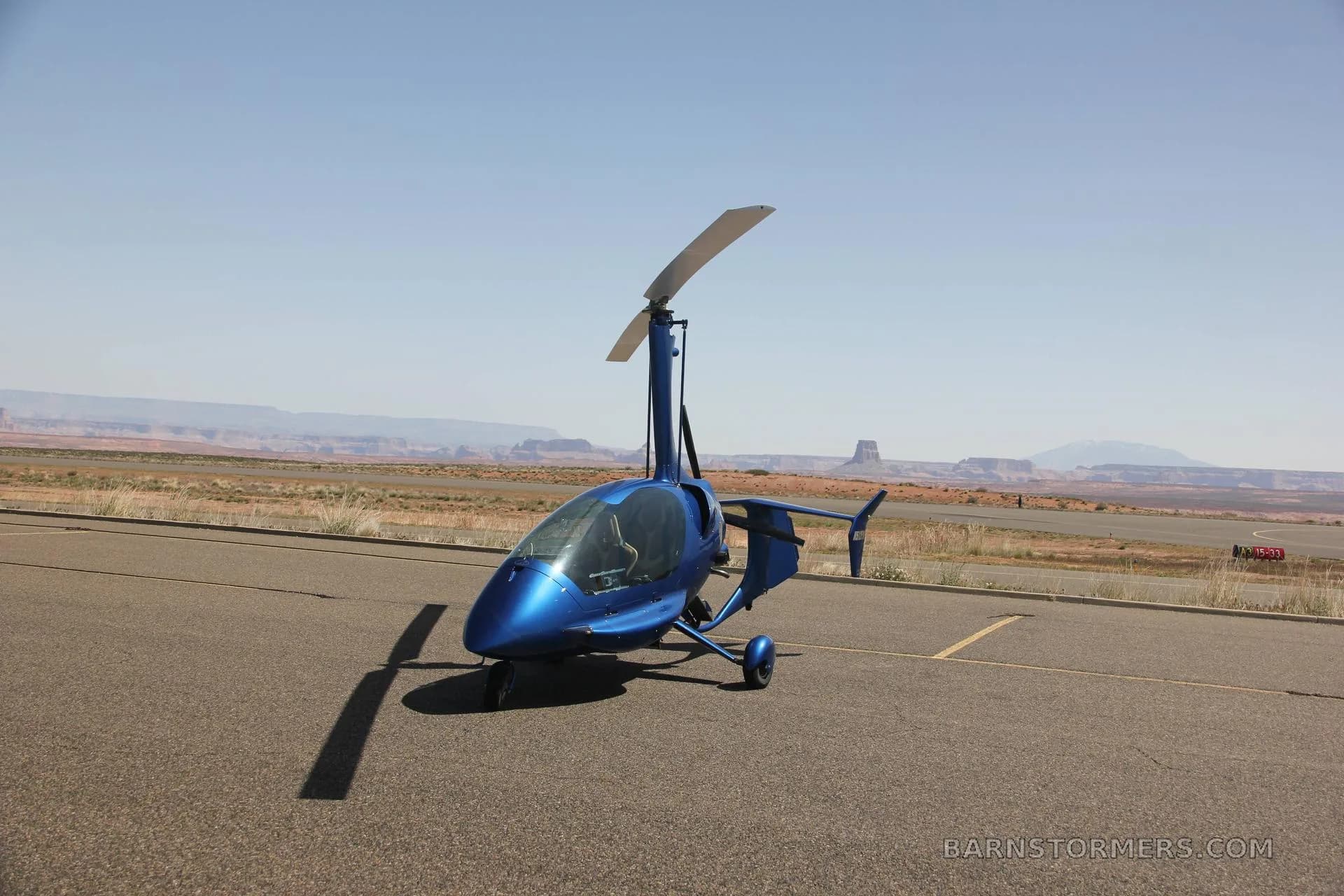 A blue enclosed gyrocopter is parked on an airfield with a vast desert landscape and mountains under a clear sky.