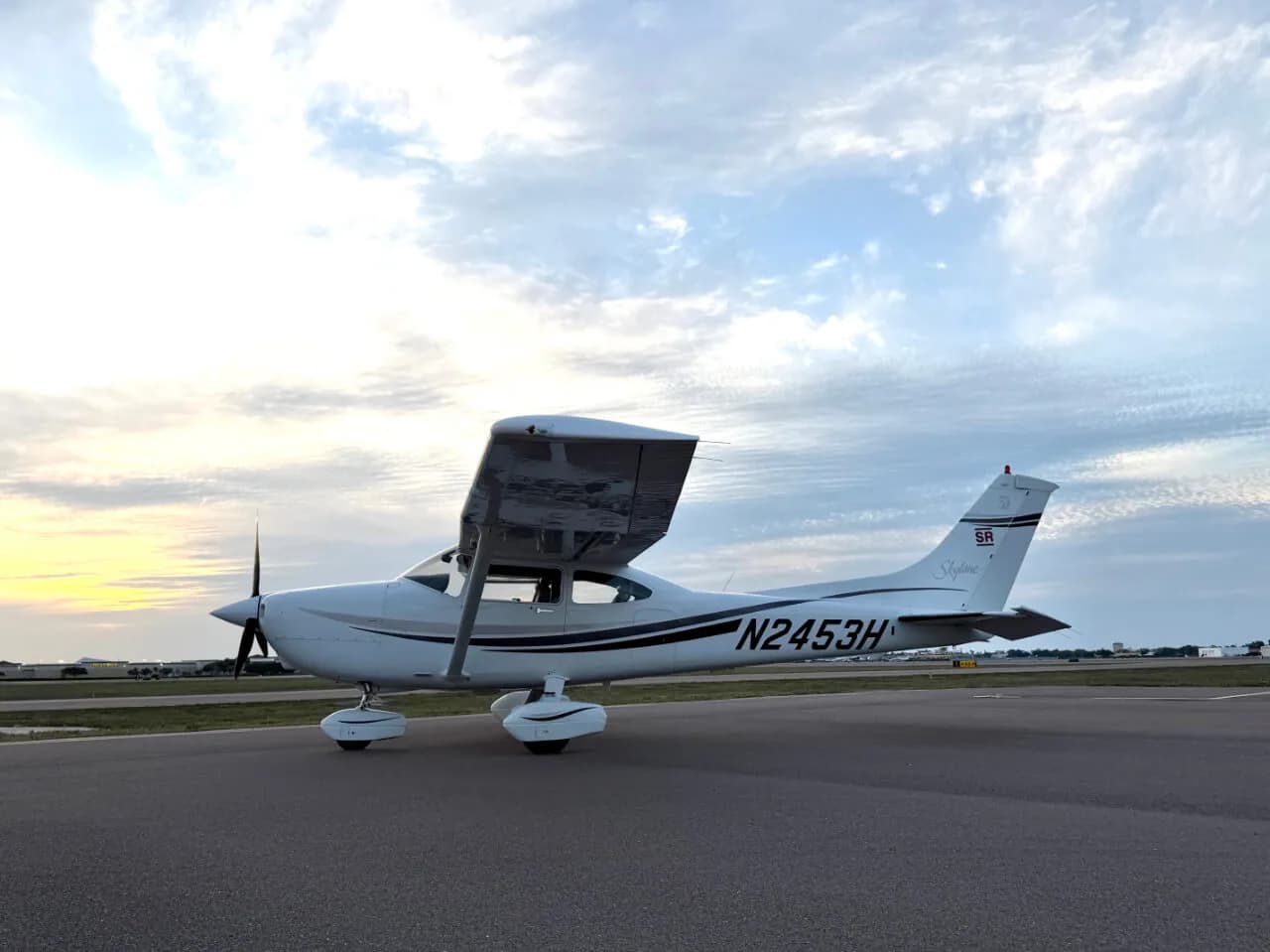 A white Cessna 182 Skylane with black stripes and registration N2453H is parked on a tarmac under a cloudy sky at sunset.