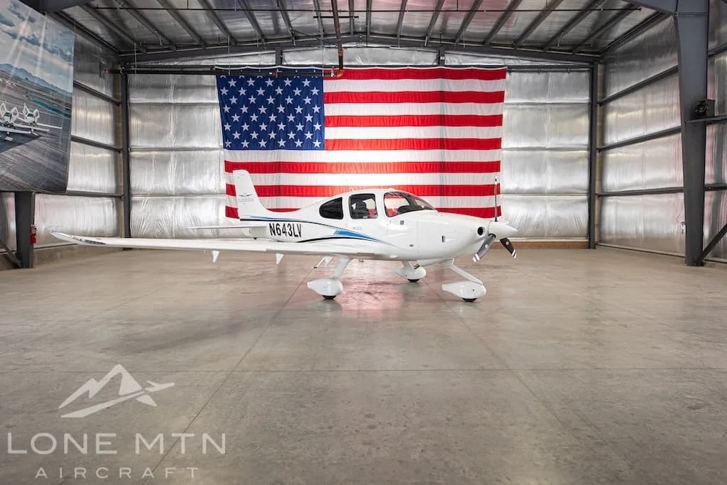 A white Cirrus SR22 aircraft with blue and black stripes, tail number N643LV, parked in a hangar with an American flag in the background.