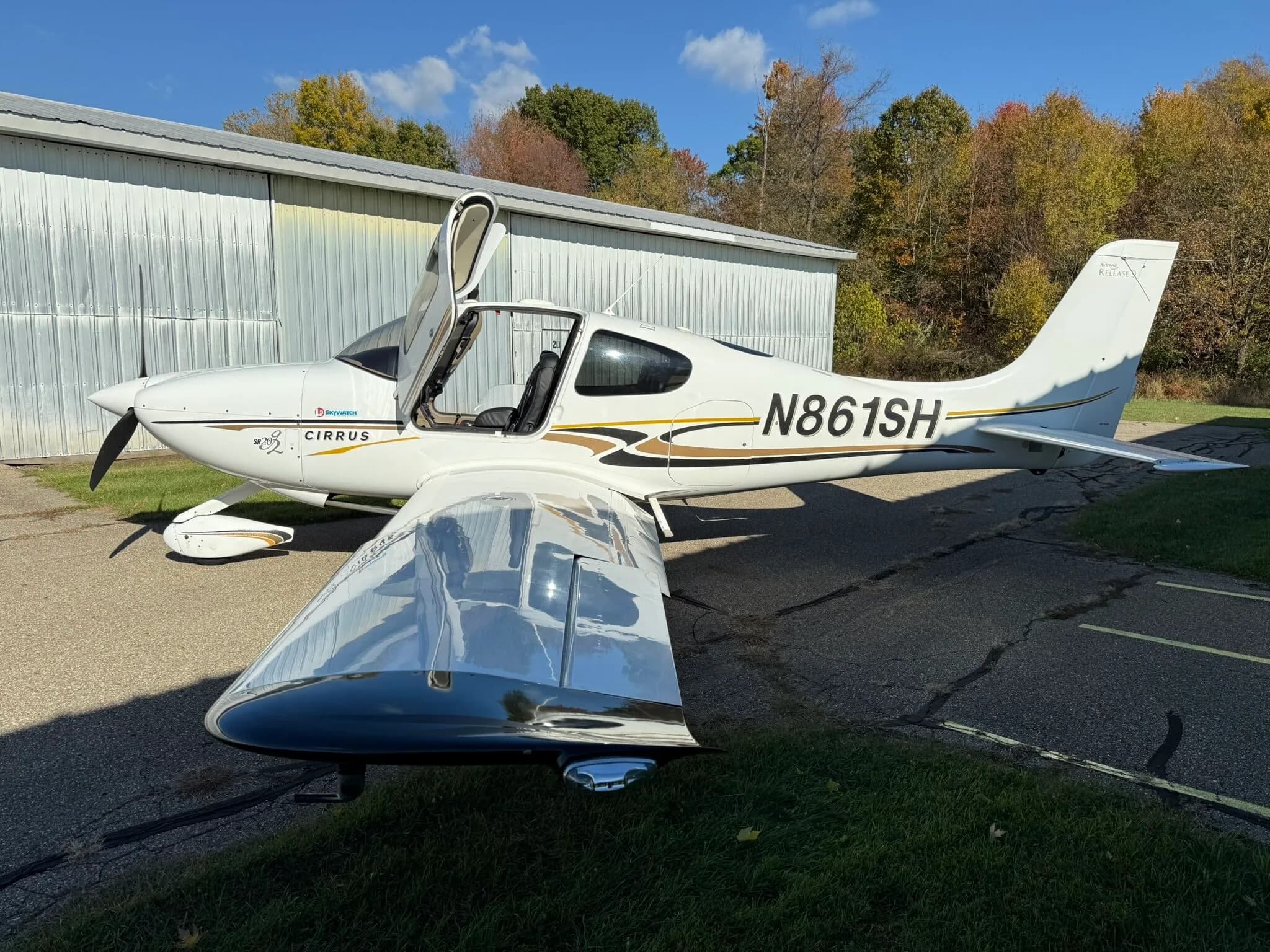 A white Cirrus SR20 aircraft with black and gold stripes, registration N861SH, parked on an asphalt ramp with its doors open.