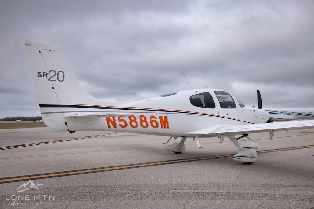 A rear-side view of a white Cirrus SR20 aircraft with orange and black stripes, registration N5886M, on an airfield.