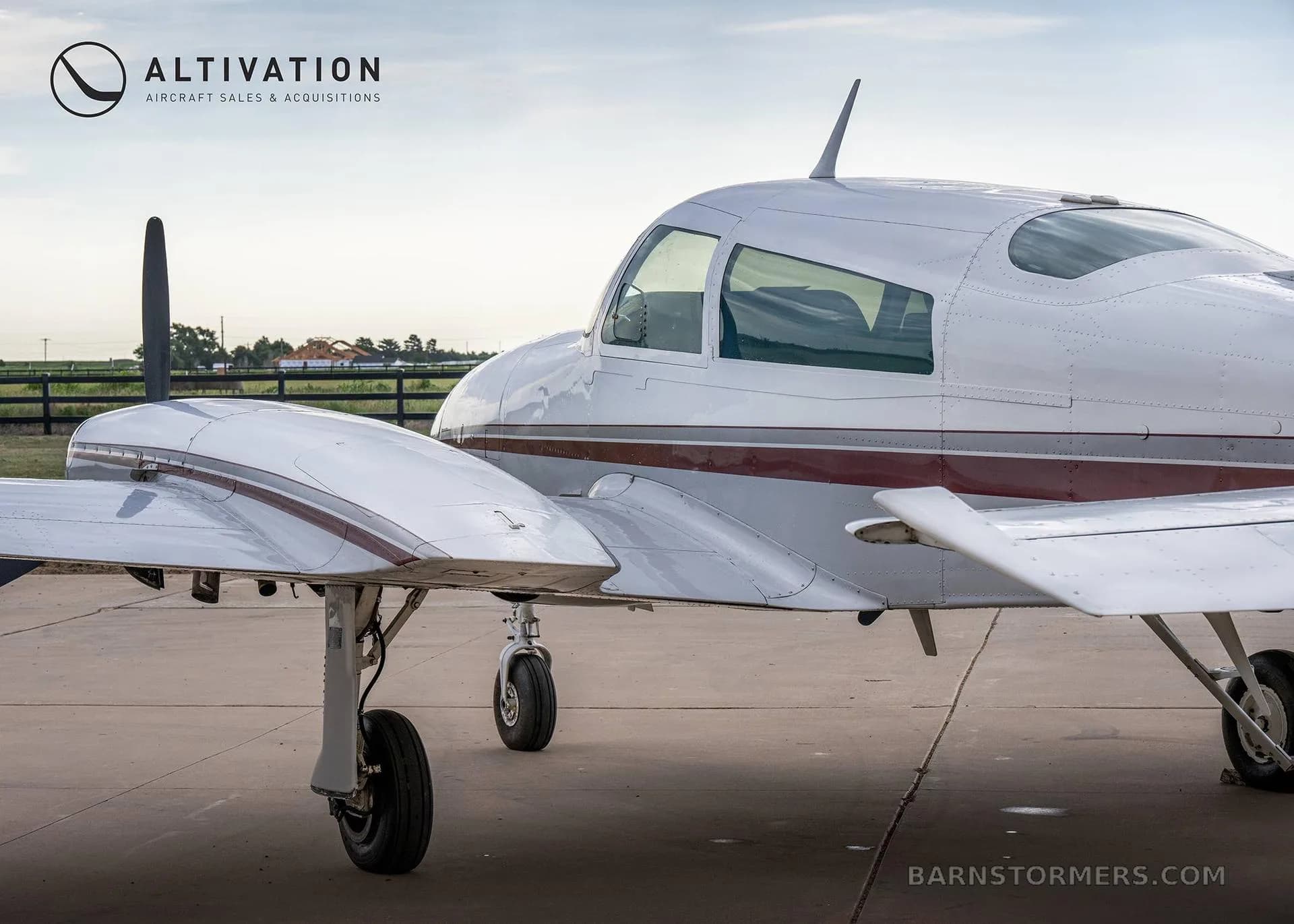 A close-up side view of the fuselage and right wing of a white twin-engine propeller aircraft with maroon stripes, parked on an airfield.