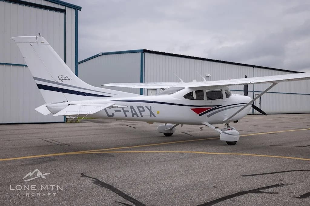 A white Cessna 182 Skylane with grey and blue stripes, registration C-FAPX, parked on an asphalt ramp with hangars in the background.