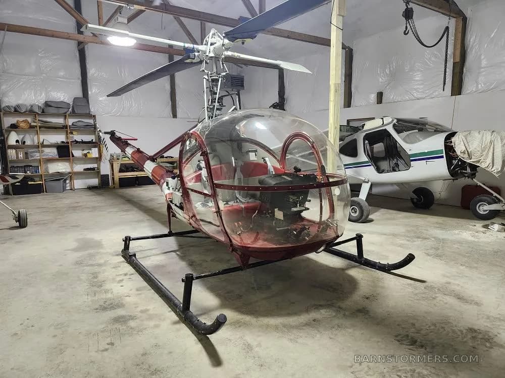 A red and white helicopter with a clear bubble canopy is parked in a hangar next to a white and green fixed-wing aircraft.