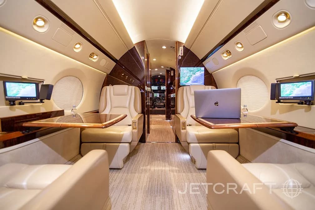 A private jet cabin with two sets of facing beige leather seats, each with a wooden table and entertainment screen.