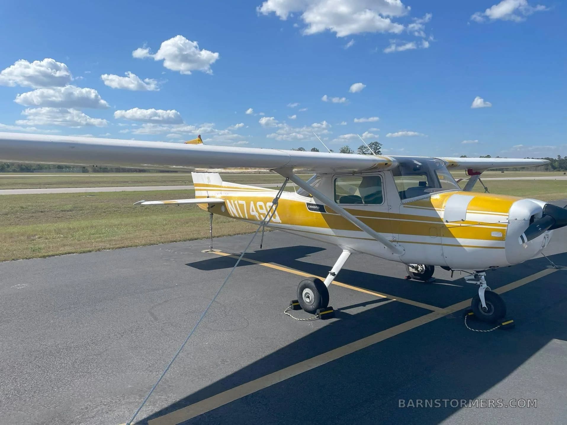 A white and yellow Cessna 172 (N1749Q) is parked on an asphalt tarmac with a grassy field and trees in the background.