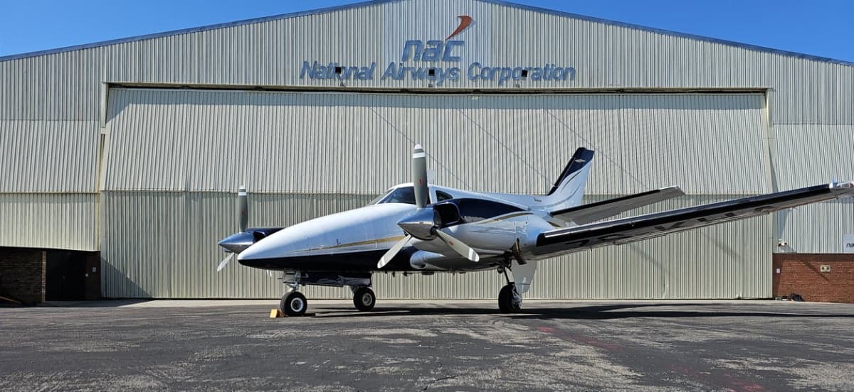 A white and black twin-engine aircraft is parked on an asphalt lot in front of a large hangar.