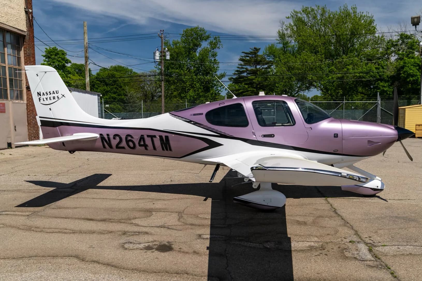 A Cirrus SR22 aircraft with a metallic lavender and white paint scheme, registration N264TM, parked on an airfield with trees in the background.