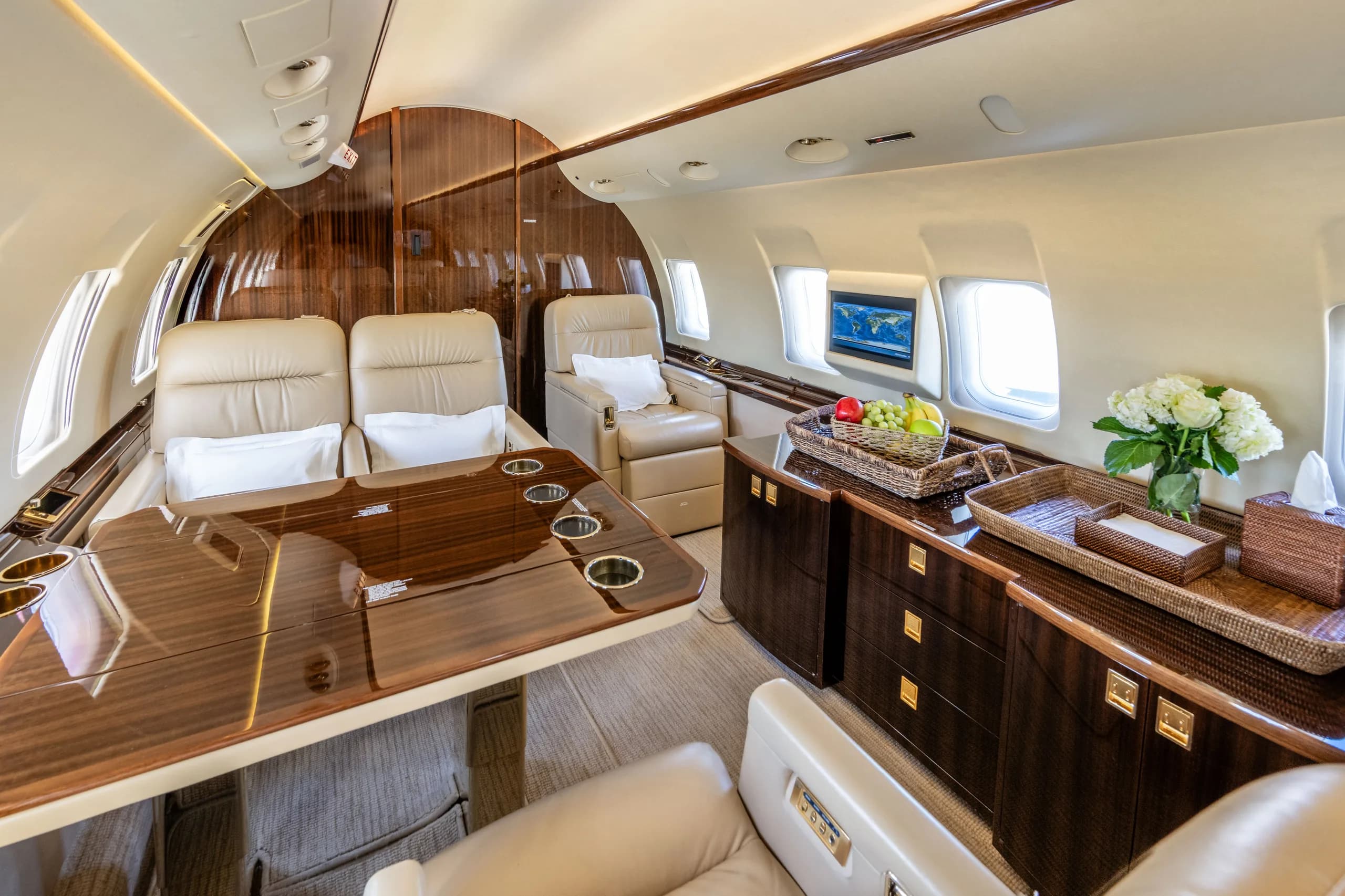 A section of a private jet cabin with a large polished wooden table, beige leather seats, and a credenza with a fruit basket and flowers.