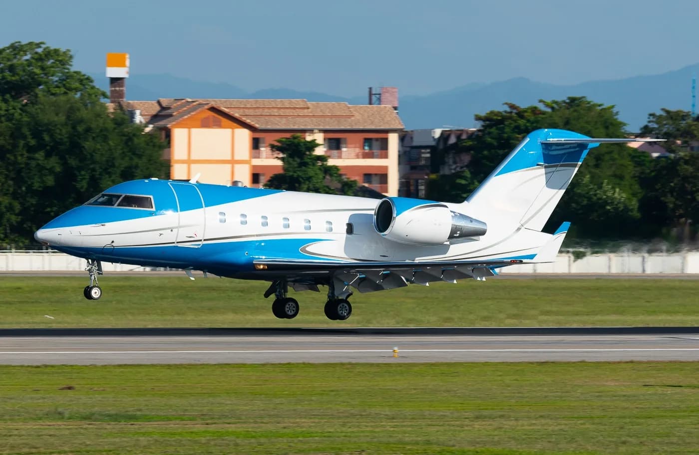 A blue and white Bombardier Challenger business jet with landing gear extended, just above a runway with buildings and trees in the background.